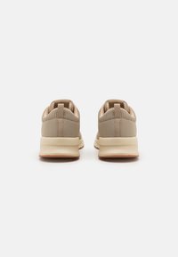 Zapatillas deportivas beige con parte superior de tela texturizada, cuello elasticado y suela de goma, que cuentan con una pestaña en la parte trasera para facilitar su uso.