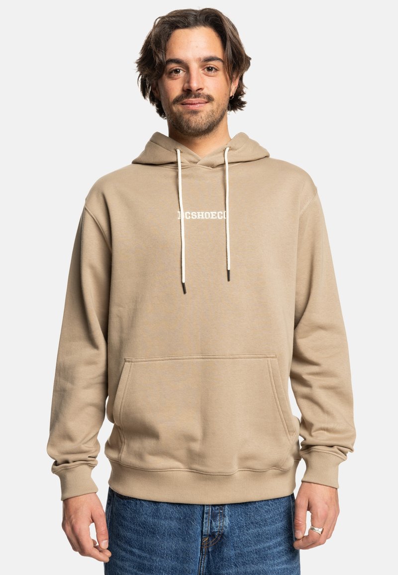 Beige sweatshirt met capuchon en voorzak, met koordjes en geborduurd logo. Zacht materiaal en relaxte pasvorm. Gecombineerd met blauwe denim jeans.