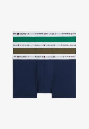 Jeune homme debout pieds nus devant un fond uni, portant un boxer vert Tommy Hilfiger avec une ceinture blanche.