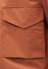 Μεγάλη ορθογώνια τσέπη από ύφασμα σε σκουριά χρώμα με μυτερή κάλυψη και εμφανείς ραφές σε ένδυμα.