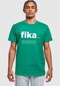 Zaļš kokvilnas T-krekls ar īsām piedurknēm, kas rotāts ar vārdu "fika" lieliem baltiem burtiem. Apakšā ir iekļauta mazāka teksta apraksts.