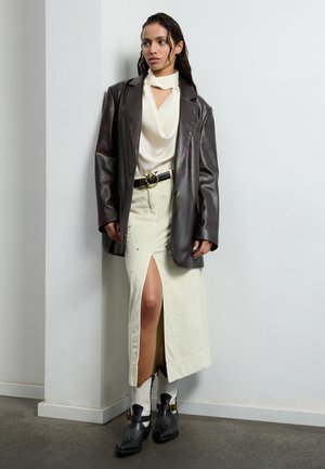 Brun läderblazer, krämfärgad silktop, ljusbeige kjol med hög slits, svart bälte med guldfärgade detaljer, svarta ankelstövlar med vita accenter.