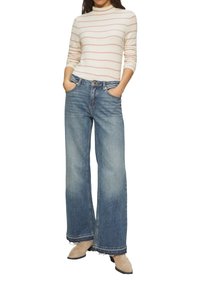 Frau in einem weißen Langarmshirt mit rosa Streifen, weit geschnittener blauer Jeans und beigen Wildlederschuhen, die die Hände in den Taschen hat.