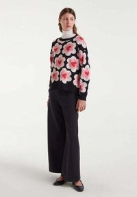 Mujer que lleva pantalones anchos negros, un suéter negro con grandes patrones florales en rosa y rojo, un cuello de cisne blanco y zapatos planos negros.