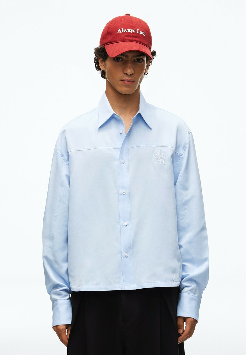 Jeune homme portant une chemise bleu clair boutonnée et une casquette rouge avec les mots "Always Late" brodés en blanc.