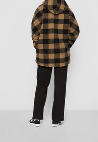 Fleecejakke i sort og tan ternet mønster med hætte, parret med sorte cargo-bukser og mørke sneakers. Teksturen ser blød og varm ud.