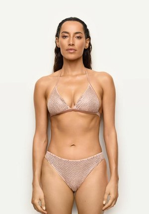 Žena s dlouhými tmavými vlasy, oblečená v světle růžových háčkovaných bikinách, stojí před jednoduchým béžovým pozadím, čelem vpřed.