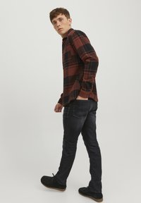 Camisa de franela a cuadros en rojo y negro, de manga larga, ajustada, combinada con jeans oscuros y zapatos de ante negros, de pie contra un fondo neutral.