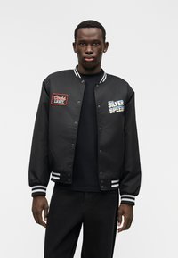 Μαύρο μπουφάν τύπου βόμβα με λευκές ρίγες στις μανσέτες και το γιακά. Διαθέτει κόκκινες ραφές με το λογότυπο "Coors Light" και μπλε ραφές με το λογότυπο "Silver Speed". Υλικό λείο και λαμπερό.