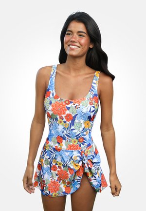 Smiling woman with long dark hair wearing a sleeveless floral swimsuit with a tied skirt detail, standing against a plain white background.