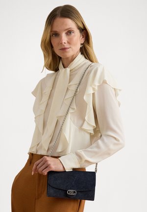 Femme portant un chemisier crème à volants et un pantalon marron, tenant un petit sac bleu foncé avec une bandoulière en chaîne argentée.