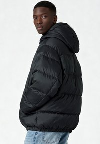 Veste matelassée noire avec capuche, comprenant des sections matelassées, une texture lisse et des poignets élastiques. Portée avec un jean denim bleu clair.