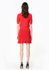 Vestito rosso lavorato a maglia con maniche corte e corpino attillato, caratterizzato da un orlo arricciato. Indossato con tacchi neri, che mettono in risalto una texture elegante.