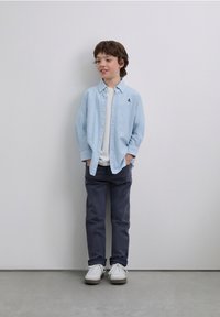 Vaaleansininen raidallinen nappipaita valkoisen t-paidan päällä, harmaat housut ja valkoiset lenkkarit; lapsi seisoo rennosti käsineen taskuissa.