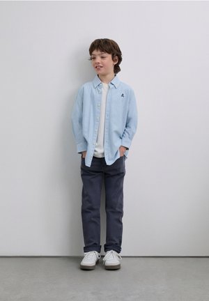 Camicia a righe azzurro chiaro sopra una maglietta bianca, pantaloni grigi e scarpe da ginnastica bianche; bambino in piedi in modo disinvolto con le mani nelle tasche.