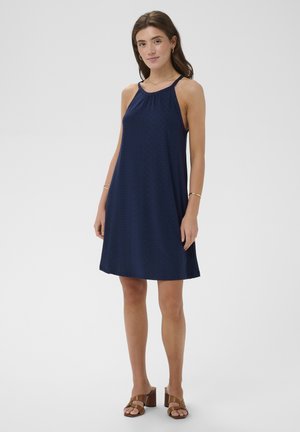 Jeune femme portant une robe bleu marine sans manches, longueur genou, avec un col licou, et des sandales à talons marron, debout sur un fond blanc.