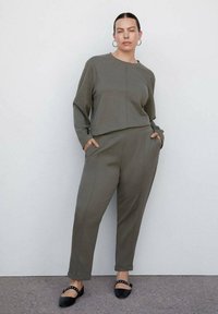 Olivengroene, lang mouwen top met een voornaaddetail, gecombineerd met bijpassende, taperende broeken met zijzakken. Zwarte platte schoenen met enkelbandjes.
