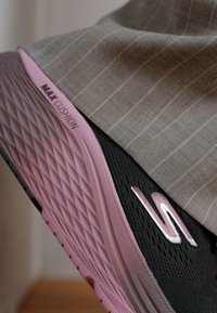 Black sneaker with textured upper and pink midsole showing "MAX CUSHION" branding; paired with a gray pinstriped fabric.