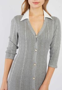 Cardigan en tricot gris avec un décolleté en V profond, des manches trois-quarts et des boutons en perles. Le tissu présente un motif texturé avec des détails en cable.