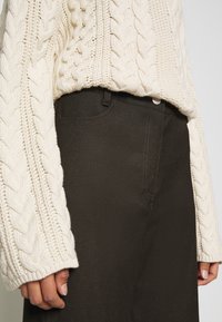 Kremowy sweter z grubym splotem i dużymi rękawami, zestawiony z ciemnobrązowymi spodniami o gładkiej fakturze i standardowych kieszeniach.