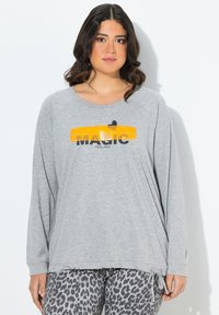 Giovane donna con capelli scuri e ricci, indossa una maglietta grigia a maniche lunghe con il testo "MAGIC FEELING" su una pennellata gialla e pantaloni con stampa leopardata.