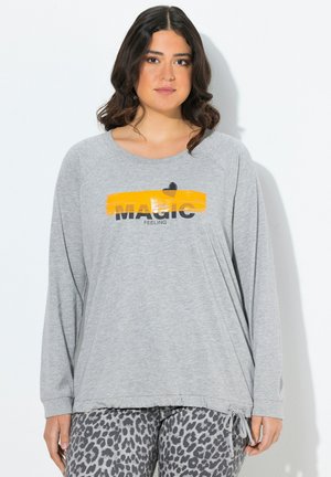 Junge Frau mit welligem, dunklem Haar, die ein graues Langarmshirt mit dem Text "MAGIC FEELING" über einem gelben Pinselstrich und einer Leopardenmusterhose trägt.