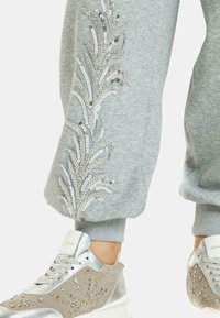 Graue Jogginghosen mit Pailletten-Blattstickerei an der Seite, kombiniert mit silbernen und beigen Sneakers, die mit dekorativen Steinen und einer Plateausohle ausgestattet sind.