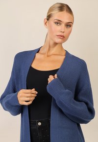 Sininen ribatattu cardigan, jossa on avoin etuosa ja pitkät hihat, valmistettu teksturoidusta kankaasta. Mallilla on päällään musta toppi ja tummat housut.