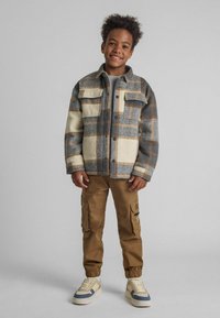Grau-beigefarbene karierte Fleecejacke mit Knopfleiste und zwei Brusttaschen, kombiniert mit braunen Cargo-Hosen und Sneakers.