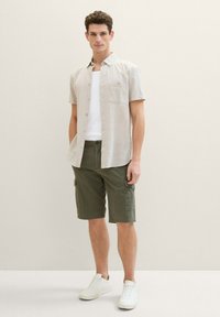 Camisa de botón sin mangas color beige con un bolsillo en el pecho, combinada con pantalones cortos cargo verde oliva y zapatillas blancas, todo sobre un fondo neutro.