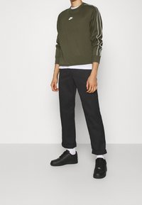 Olive green sweatshirt with white stripes, short sleeves, and logo. Black pants, cuffed at the ankles, with black sneakers and white socks.