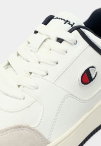 Λευκό δερμάτινο αθλητικό παπούτσι με γκρι σουέντ στοιχεία, υφασμάτινα πάνελ, λευκά κορδόνια και κόκκινο-μαύρο λογότυπο στο πλάι. Μαύρη εξωτερική σόλα.