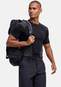 Chemise de sport noire avec panneaux texturés, associée à un pantalon foncé et un sac à dos noir. Logo visible sur la chemise et bretelles ajustables sur le sac à dos.