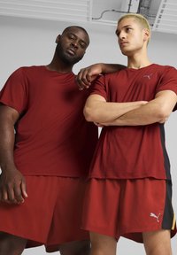 Dos modelos llevan pantalones cortos deportivos rojos y camisetas rojas a juego que presentan el logo de Puma en colores contrastantes. El diseño es deportivo y funcional.