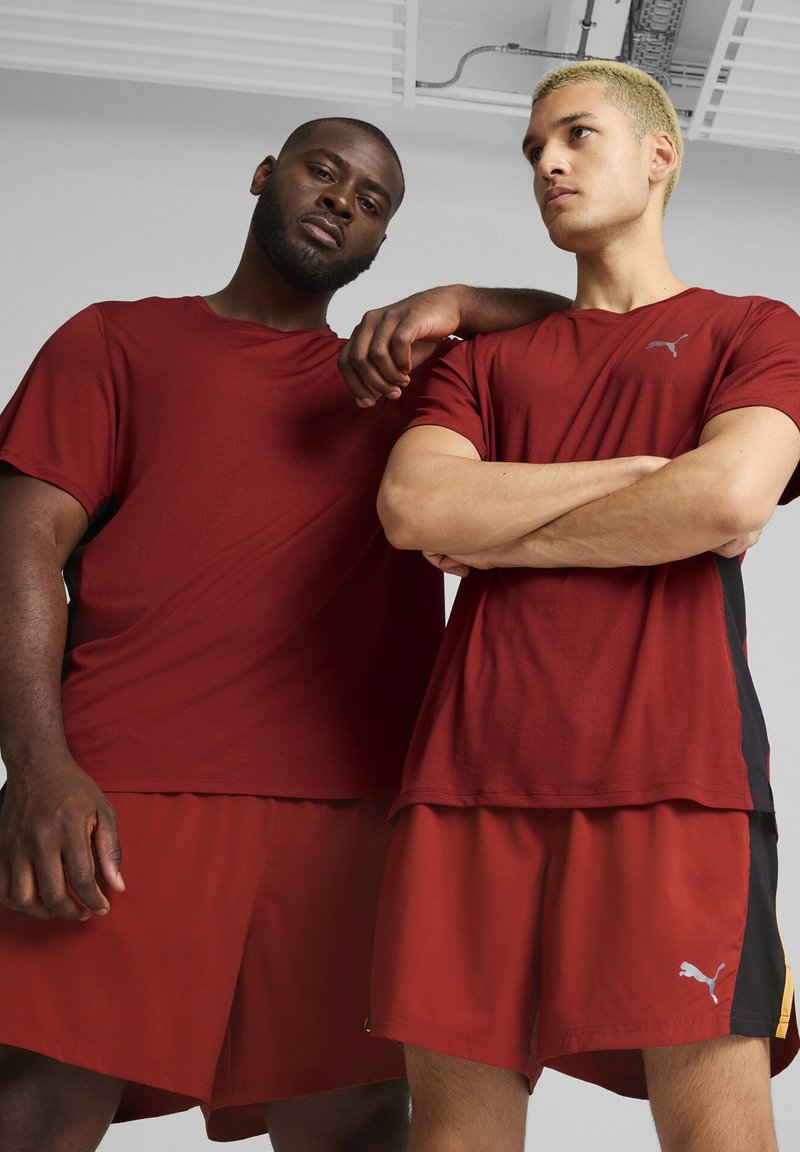Dos modelos llevan pantalones cortos deportivos rojos y camisetas rojas a juego que presentan el logo de Puma en colores contrastantes. El diseño es deportivo y funcional.