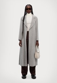 Lang grå frakk med notched krage, båret over en hvit turtleneck. Mørkebrune bukser, sorte platå sko og en lys beige håndveske.