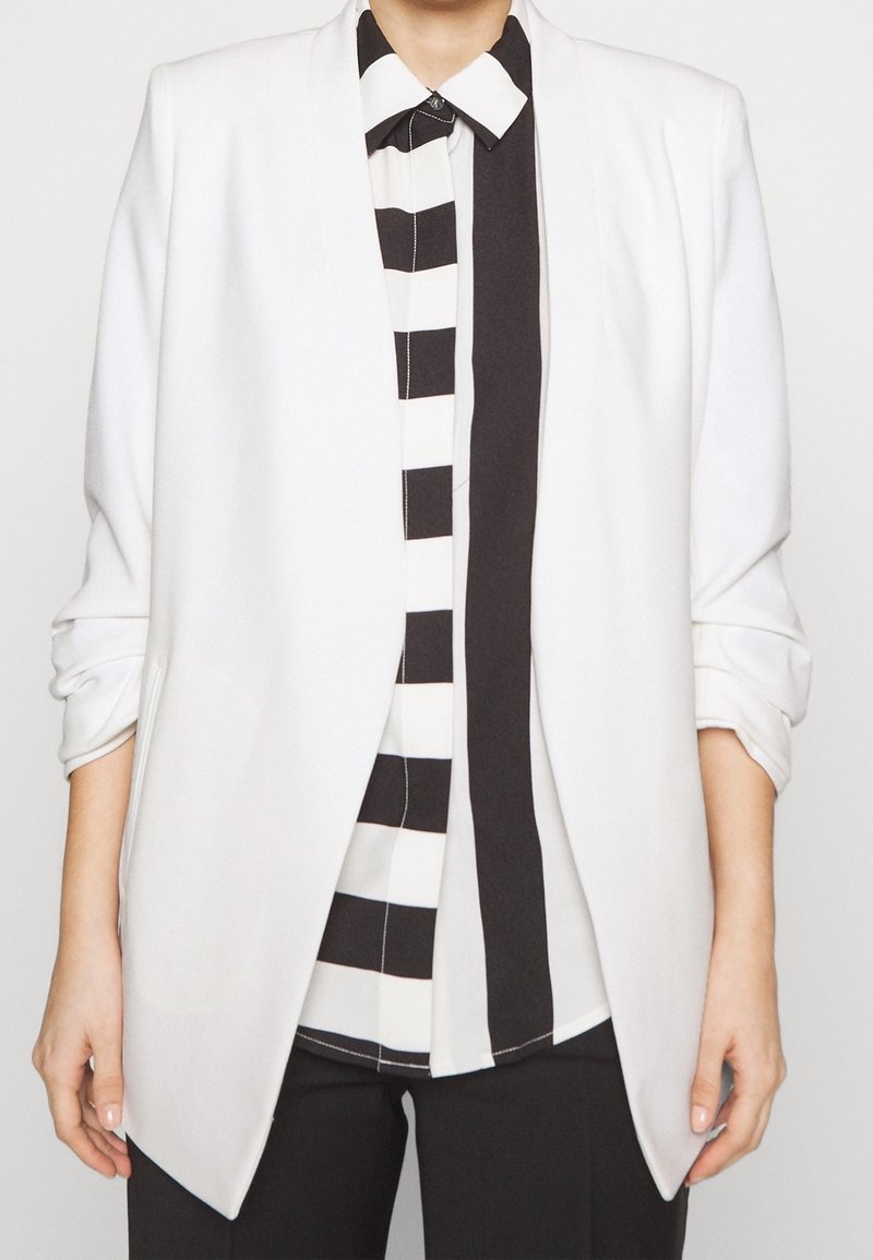 Personne portant un blazer blanc par-dessus une chemise à boutons rayée verticalement en noir et blanc, avec les manches du blazer légèrement froissées.