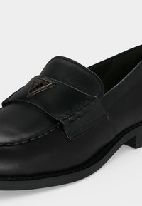 Mocassin en cuir noir avec un talon bas, un bout arrondi, des détails cousus et un logo Guess triangulaire en métal sur la bride supérieure.