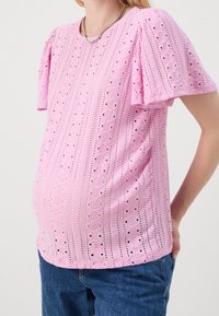 Różowa bluzka z krótkim rękawem o teksturowanym wzorze z oczkami, z dekoracyjnymi otworami i luźnym krojem, zestawiona z niebieskimi dżinsami.