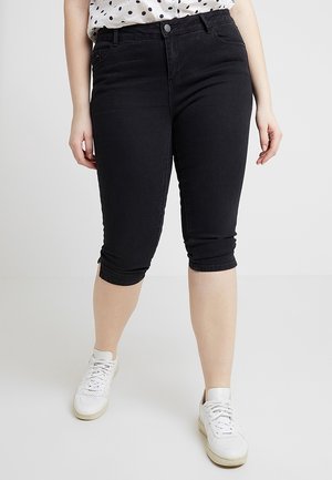 Kvinde iført sorte cropped skinny bukser med frontknap og hvide sneakers, stående på en ensfarvet baggrund.