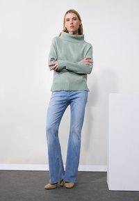 Light green, high-neck sweater with long sleeves, paired with light blue flared jeans. The model wears beige heeled shoes. Minimal background.