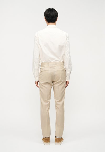 Άνδρας που στέκεται γυρισμένος με την πλάτη, φορώντας λευκό πουκάμισο μακρυμάνικο, μπεζ παντελόνι και καφέ σουέντ αθλητικά παπούτσια, μπροστά σε λευκό φόντο.