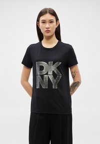 Jovem com cabelo longo e escuro, vestindo uma t-shirt preta da DKNY e calças pretas, em pé com as mãos atrás das costas contra um fundo simples.