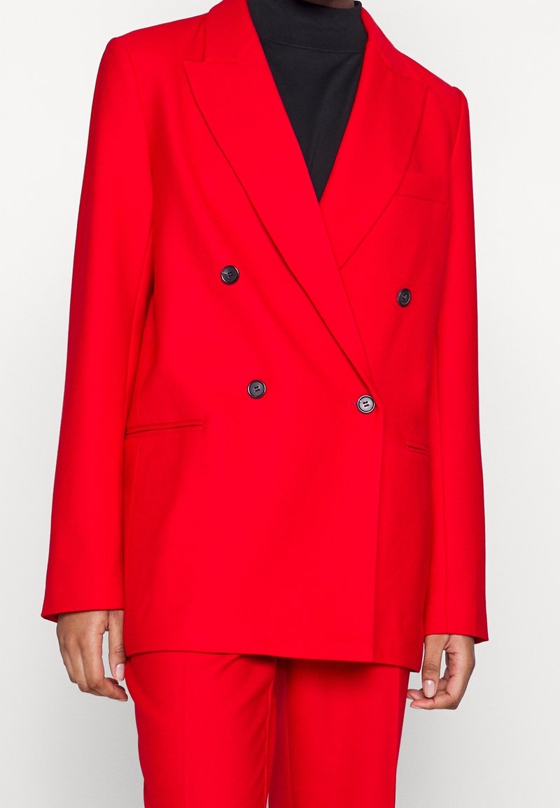 Blazer rojo de doble botonadura, hecho de una tela suave, con botones negros, un bolsillo en el pecho y solapas estructuradas.