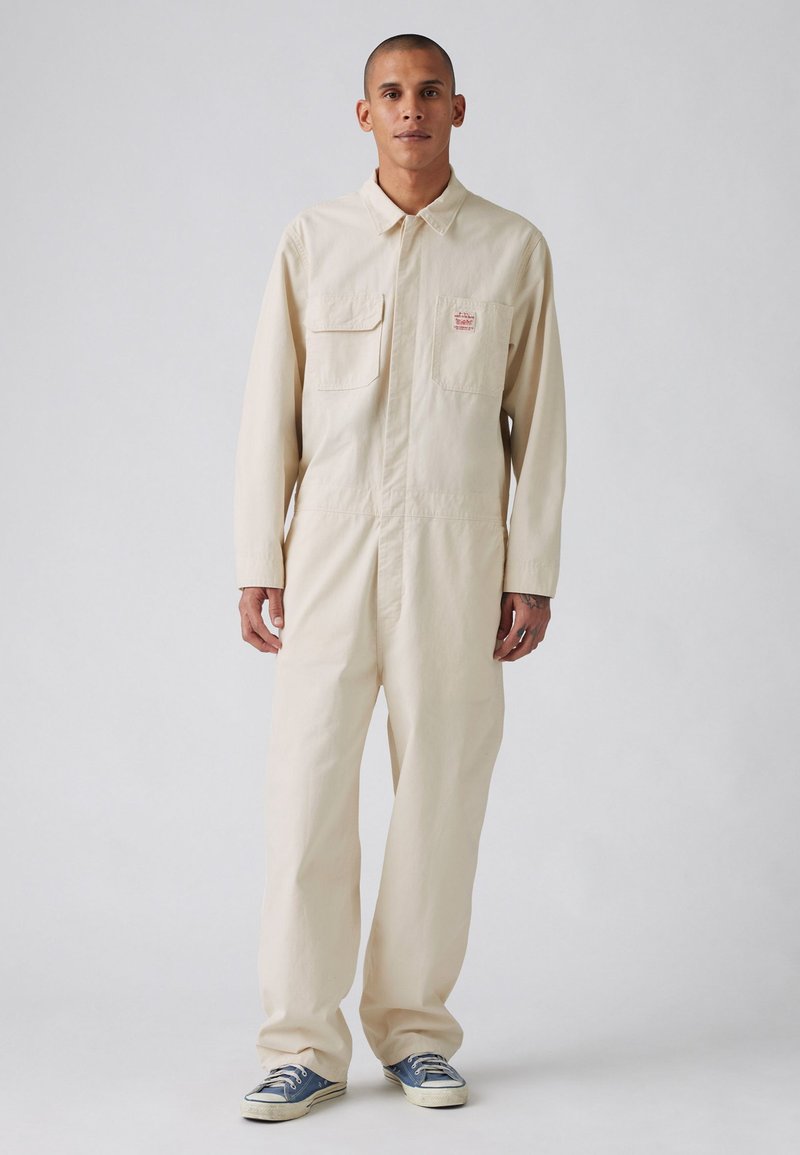 Combinaisons en coton beige avec manches longues, deux poches poitrine et coupe décontractée, associées à des chaussures en toile bleue. Design et texture simples.