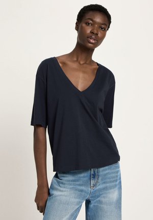 Woman wearing a loose dark navy V-neck shirt and light blue jeans, standing against a plain light background.