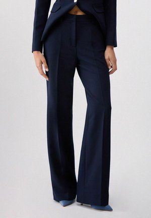 Persona che indossa pantaloni ampi a vita alta blu navy con blazer abbinato e tacchi a punta azzurri, in piedi davanti a uno sfondo semplice.