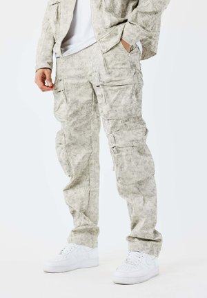 Homme portant un pantalon cargo gris clair à motifs, une veste assortie, des baskets blanches et une chemise blanche, debout avec une main dans la poche.