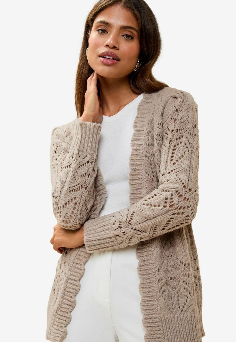 Cárdigan de punto en un suave beige con patrón de calado, con puños y dobladillo acanalados, llevado sobre una blusa blanca, que presenta mangas largas y un corte relajado.