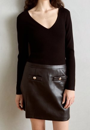 Kvinde iført sort ribbet V-hals top med lange ærmer og en sort læder mininederdel med forlommer og guldfarvede knapdetaljer.